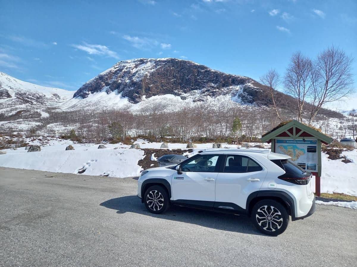 A white small SUV parked on the side of the road with a snow-covered mountain in the background